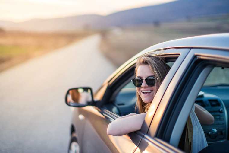 A female with sunglasses pokes her head out of a car window and smiles